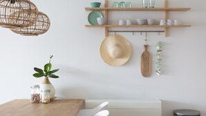 Un comedor con mesa de madera, sillas blancas, un sombrero colgando y una estantería con copas y cuencos.