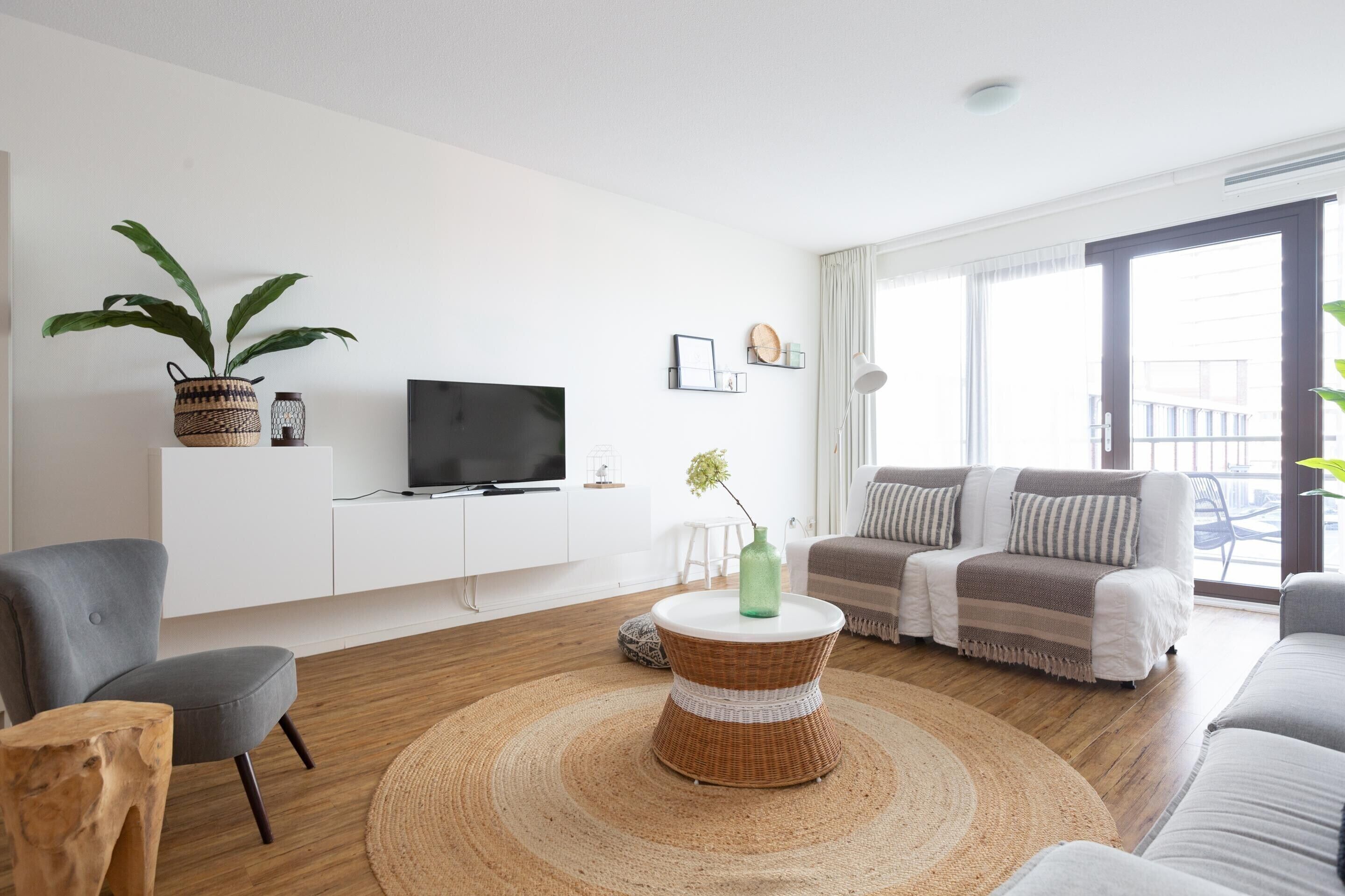 Uma sala de estar moderna com uma TV de tela plana, uma mesa de centro de vime, uma poltrona cinza, um sofá e uma grande janela com vista.