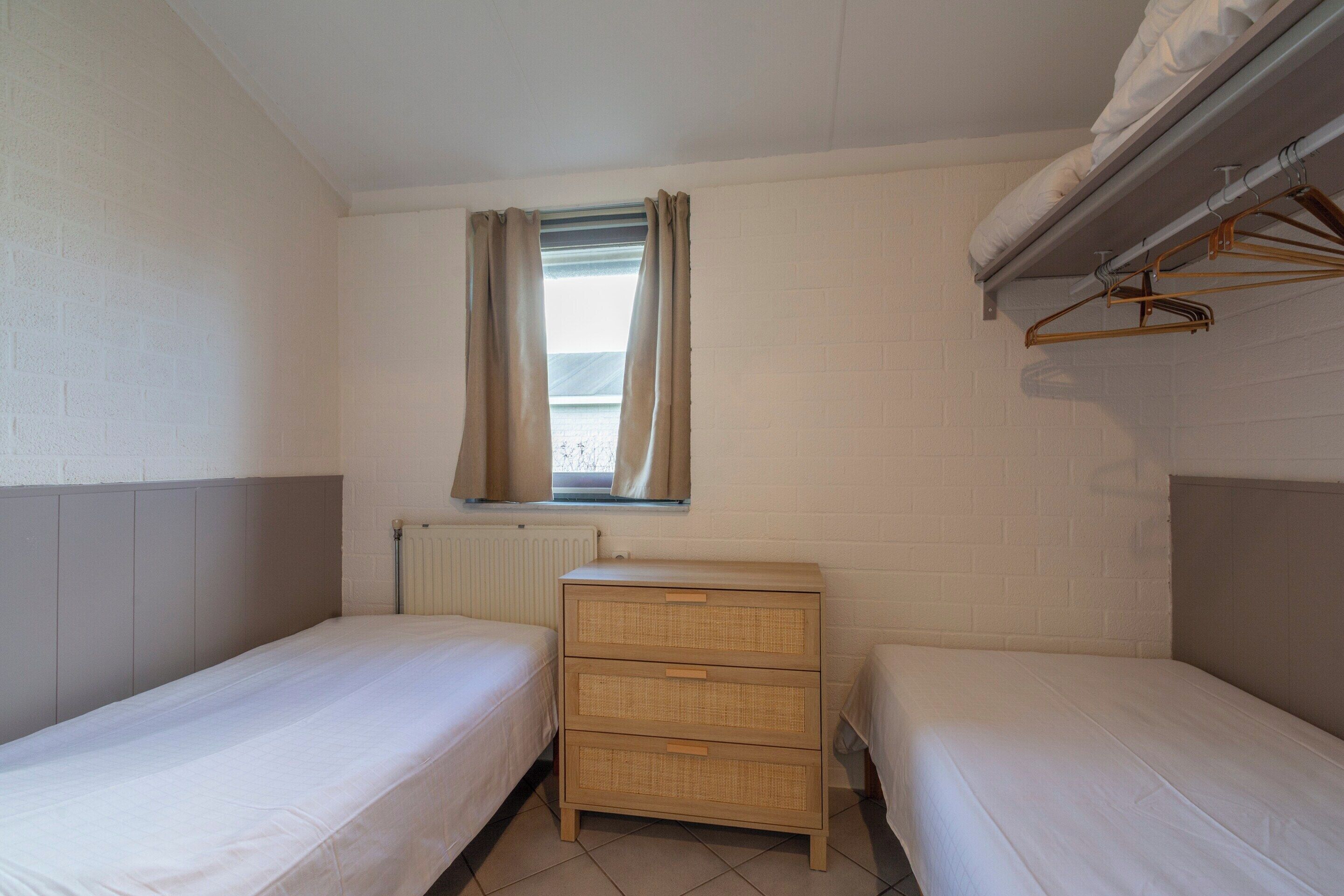 Quarto pequeno e limpo com duas camas individuais, cômoda de madeira, radiador e janela com cortinas.