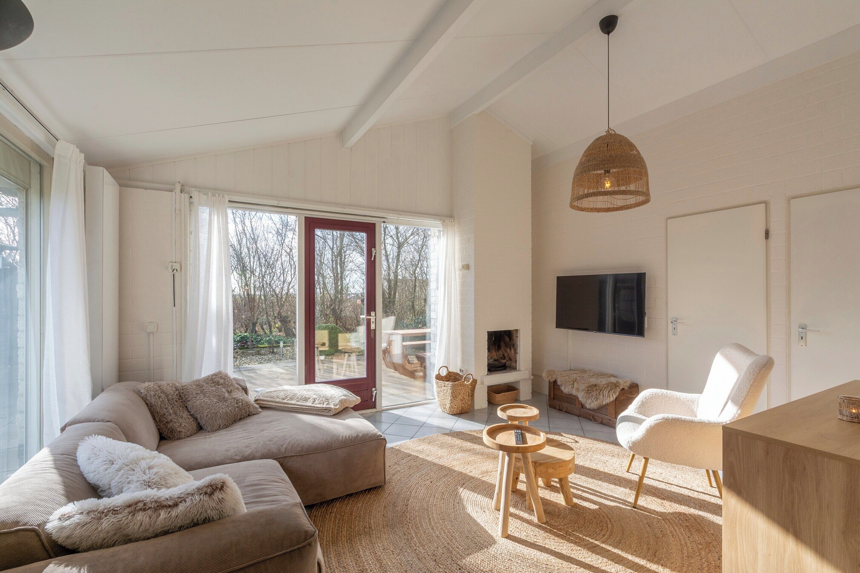 A spacious living room with a sofa, armchair, and a TV. There is a fireplace, a dining table, and a large window with curtains.