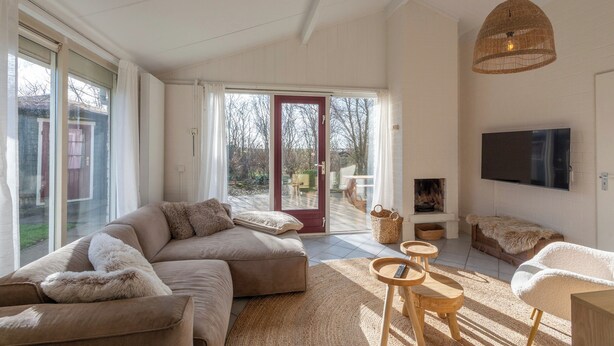 A spacious living room with a large sectional sofa, a fireplace, a TV, and a large window with a sliding glass door leading to an outdoor deck. - Holiday Home in Park De Watersnip Near Beach (Petten)