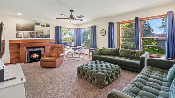 Ein Wohnzimmer mit Kamin, Ledersessel, grünem Sofa, Hocker, Esstisch und blauen Vorhängen.