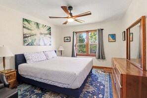 Un dormitorio con una cama, un ventilador de techo, una ventana con cortinas y un cuadro grande en la pared.