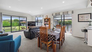 A spacious living room with a large TV, a dining table with chairs, a sofa, and a bookshelf. - Rangi Marie - Alexandra Holiday Home (Alexandra)