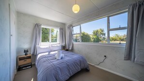 Een slaapkamer met een bed, nachtkastje, lamp, raam met gordijnen en uitzicht op groen buiten.