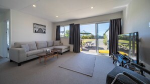 Een woonkamer met een bank, salontafel en televisie. Er is een schuifdeur die leidt naar een buitenruimte met een tafel en stoelen.