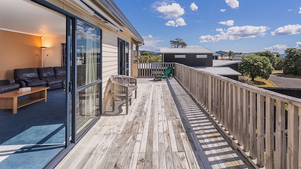 Ein modernes Haus mit einer Holterrasse, einer Glastür, die in einen Wohnbereich führt, und Blick auf die umliegende Landschaft.