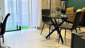 Een moderne woonkamer met een eethoek, met een glazen tafel, zwarte stoelen, een bank met gele kussens en een groot raam met dunne gordijnen.