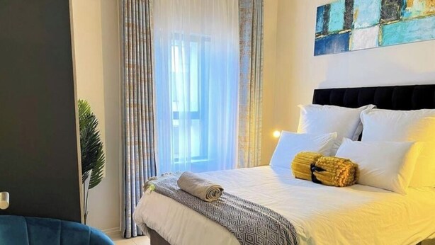 Een net opgemaakt bed met witte lakens en twee opgerolde handdoeken, een blauwe fauteuil en een groot raam met dunne gordijnen.