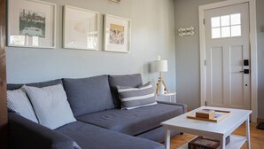 Un soggiorno moderno con un divano grigio, un tavolino bianco e una libreria. Si notano tre quadri appesi al muro e una porta con una finestra.