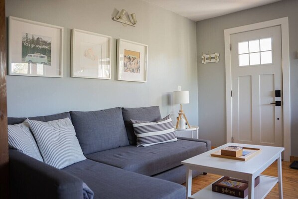 A modern living room with a grey sofa, a white coffee table, and a bookshelf. There are three framed pictures on the wall and a door with a window. - Coastal Suite – Waterfront Harbour Retreat in the Heart of Ucluelet (Ucluelet)