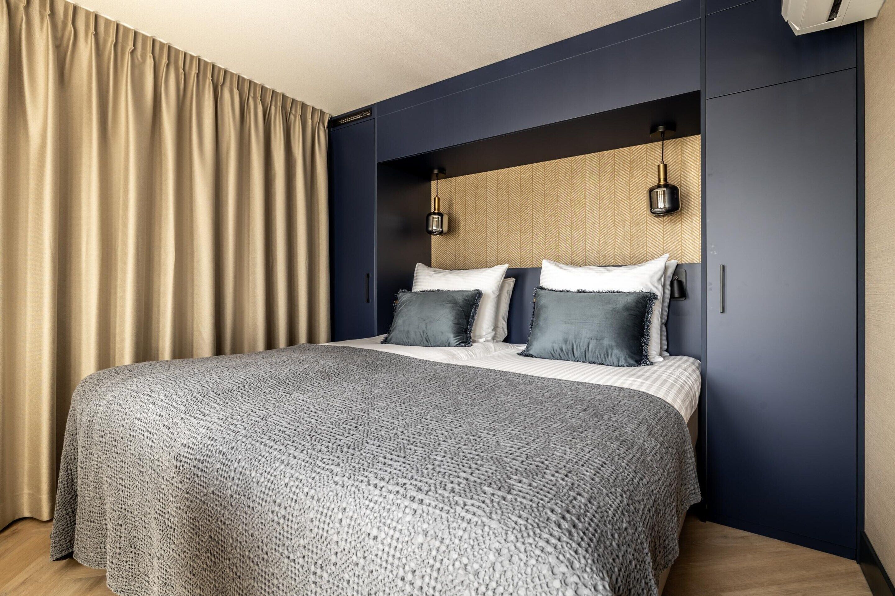 A modern hotel room with a large bed, dark blue walls, and a wooden headboard.