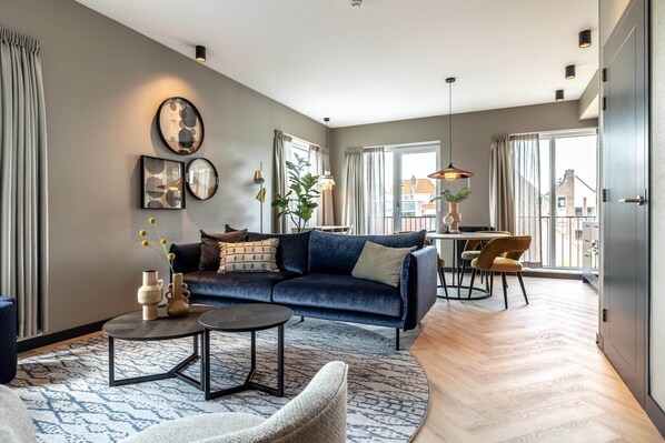 A modern living room with a dark blue sofa, a round coffee table, and a patterned rug. There is a dining area with a table and chairs, and a large window with curtains. - Cozy Apartment in the Center of Delft (Delft)