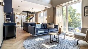 Ein modernes Wohnzimmer mit einem blauen Sofa, einem Couchtisch und Blick ins Freie durch große Fenster.