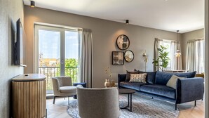 Ein modernes Wohnzimmer mit Sofa, Sessel, Couchtisch, großen Fenstern und einem flachbildfernseher an der Wand.