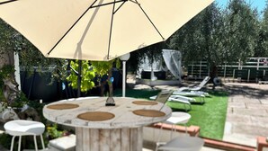 A round outdoor table with a beige umbrella, surrounded by white chairs, set on a patio with a green lawn and lounge chairs in the background.