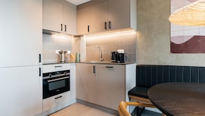 A modern kitchen with white cabinets, a built-in oven, and a coffee maker. There is a black bench and a round table in the dining area. - 1 Bedroom Apartment in Den Haag (The Hague)