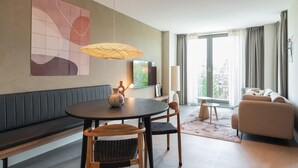 A modern living room with a round dining table, two chairs, a bench, a sofa, a TV, and a large window with curtains. - 1 Bedroom Apartment in Den Haag (The Hague)