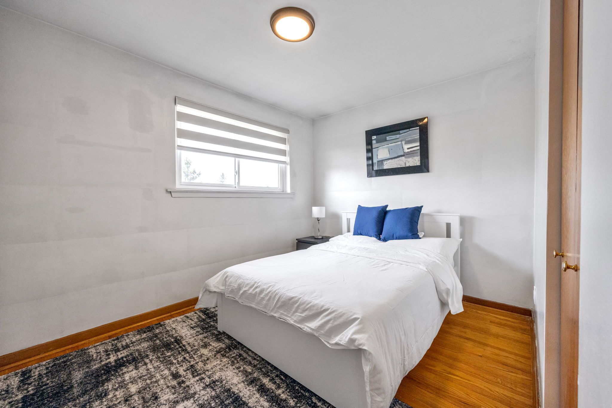 Un dormitorio con una cama, una ventana, una mesita de noche y un cuadro enmarcado en la pared.