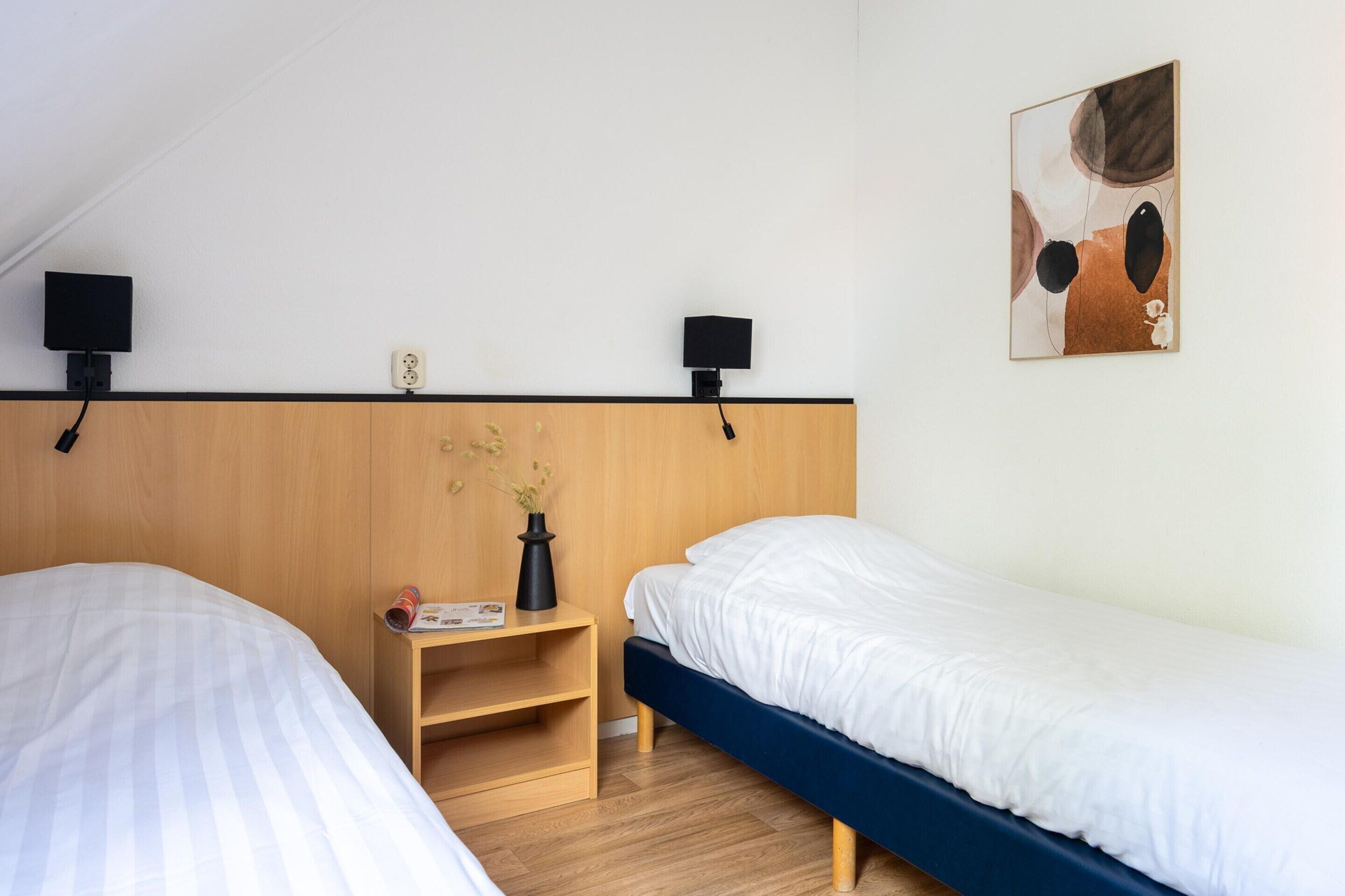 Un dormitorio con dos camas, cabecero de madera, una mesita de noche con un florero y una revista, y un cuadro en la pared.