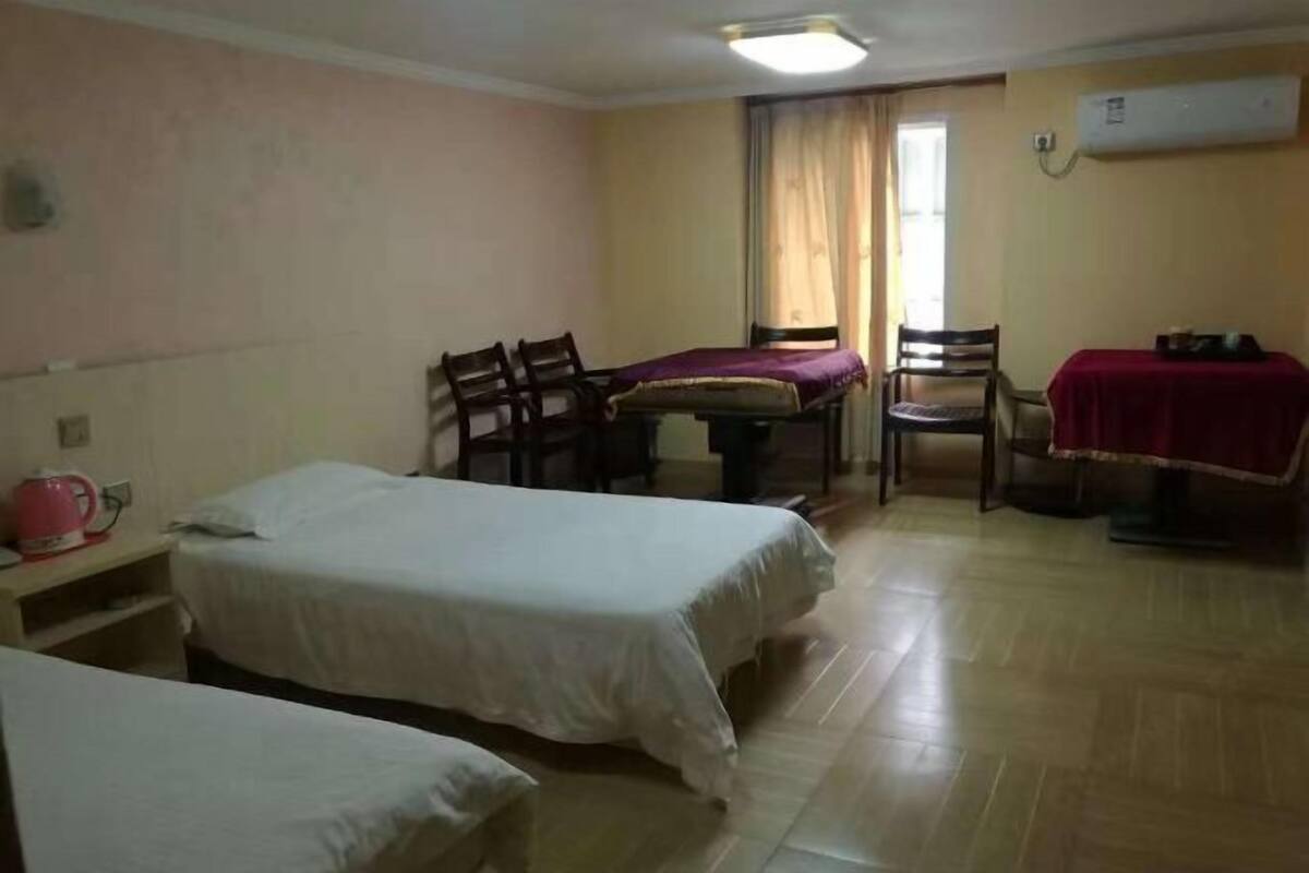 A hotel room with two beds, a dining table with chairs, a small table with a red tablecloth, and an air conditioning unit on the wall.