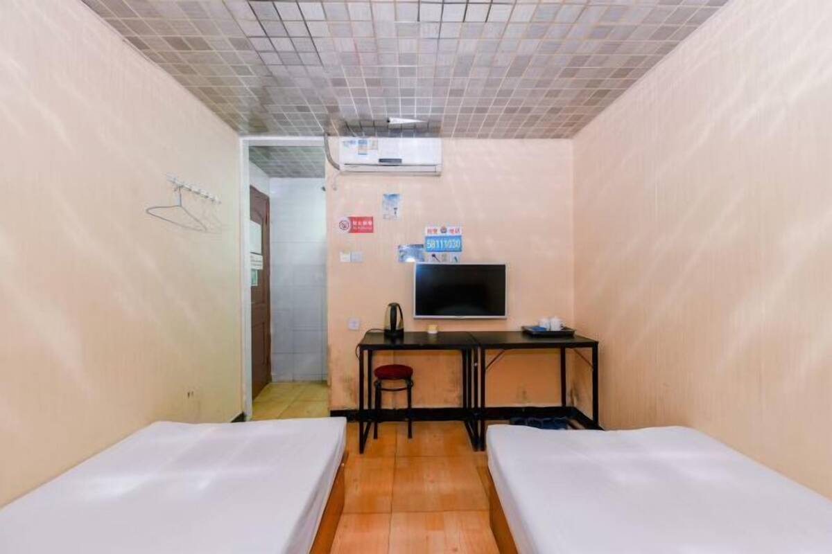 A small, single-room hotel room with a tiled ceiling, a desk with a chair, a television, and two beds.