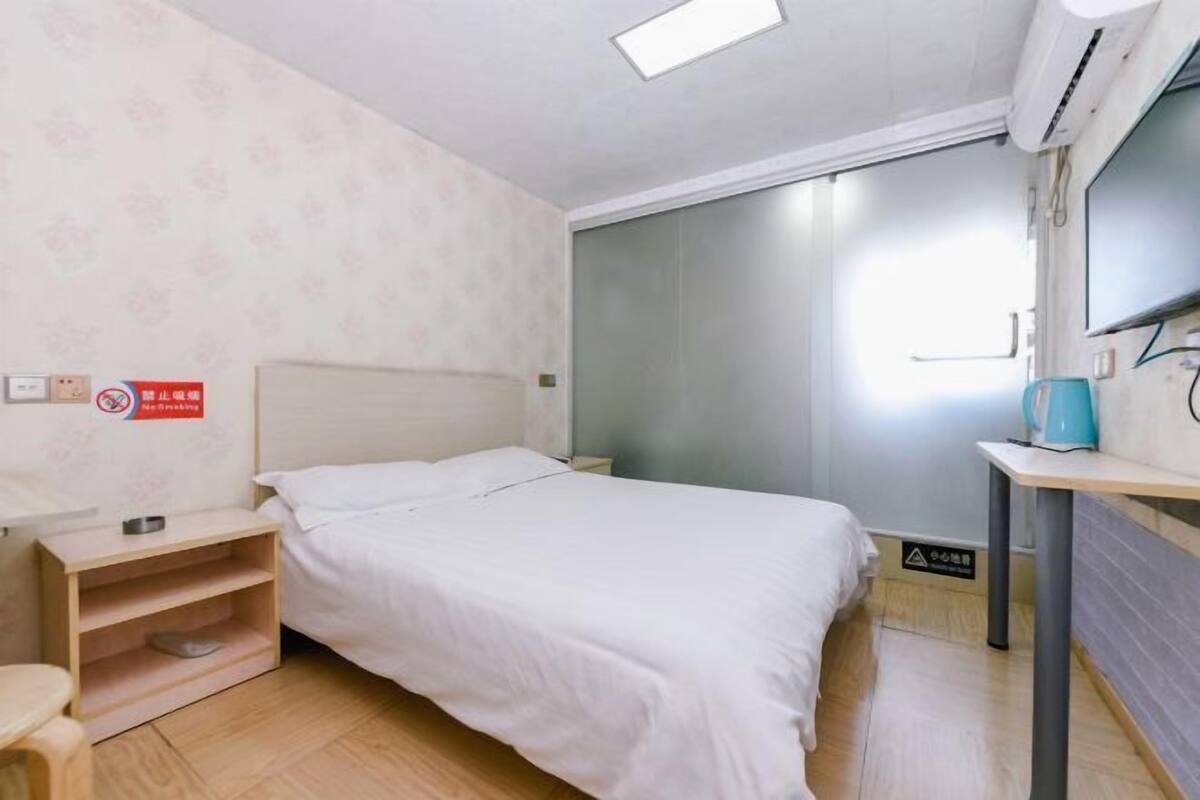 A single bed with a headboard, a bedside table, a small shelf, a wall-mounted air conditioner, and a window with a view.