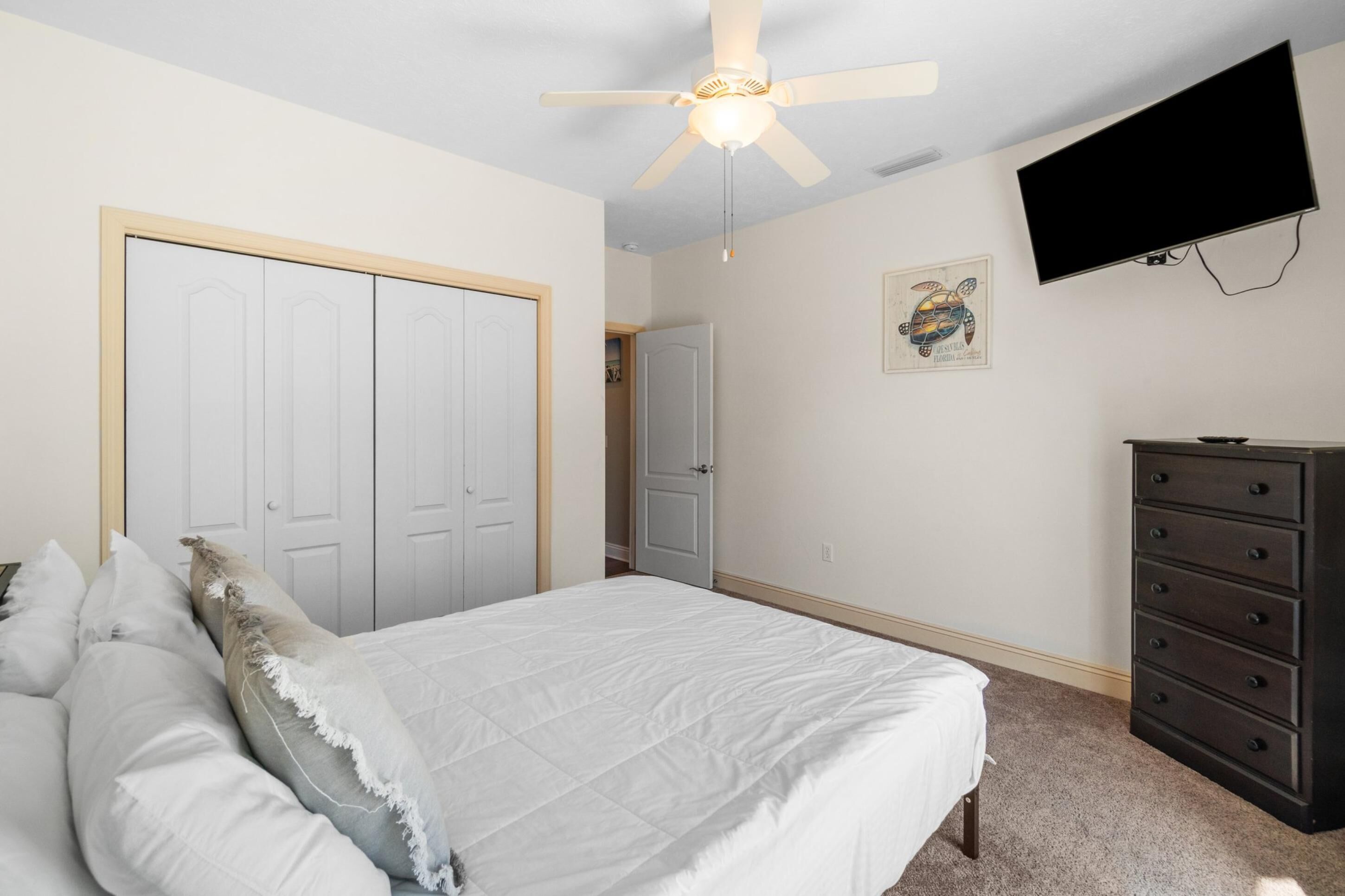 A bedroom with a bed, a dresser, a ceiling fan, a TV, and a door.