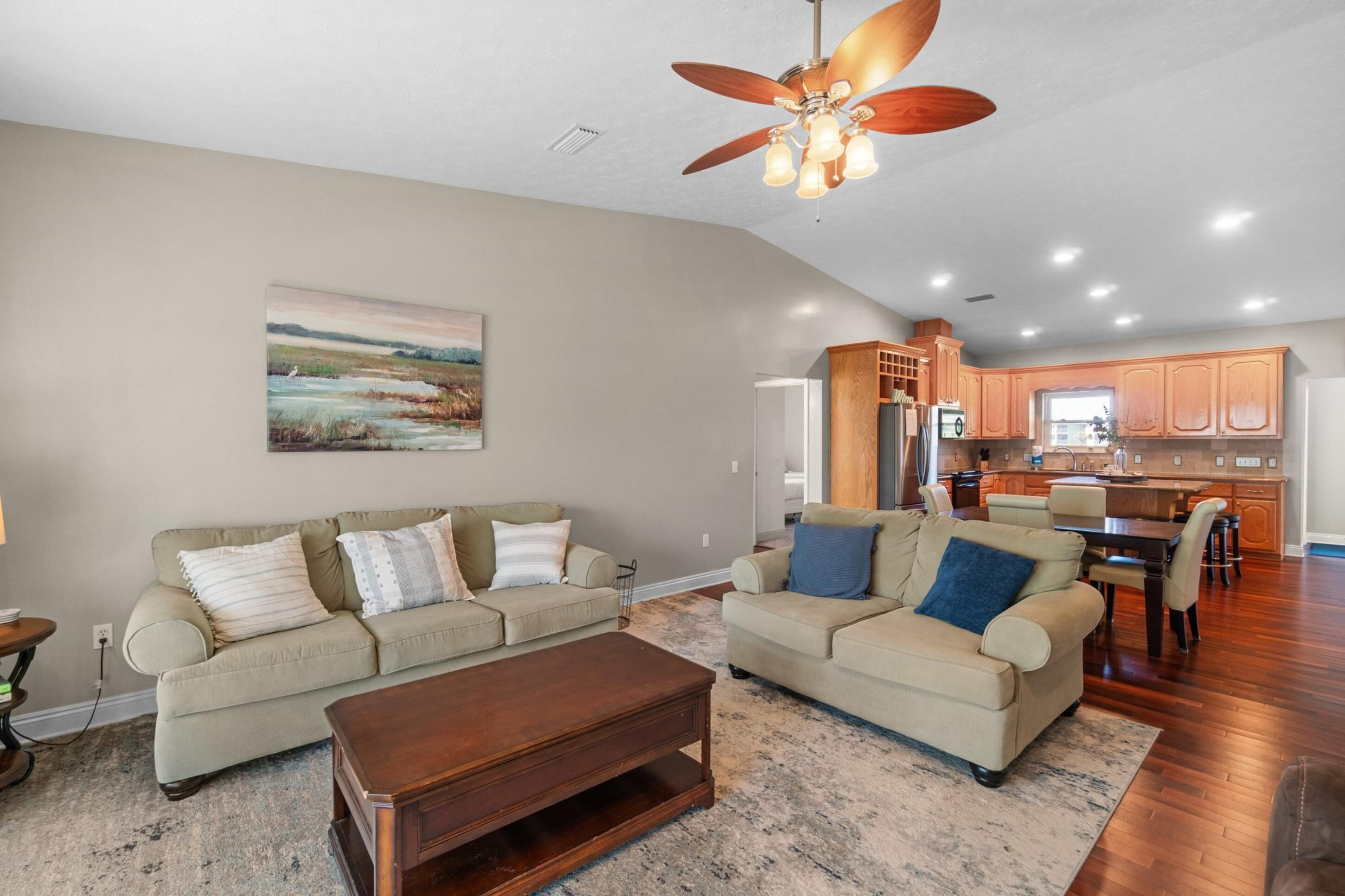 A spacious living room with two beige sofas, a wooden coffee table, and a ceiling fan. A painting adorns the wall, and the room leads to a kitchen with wooden cabinets and a dining area.