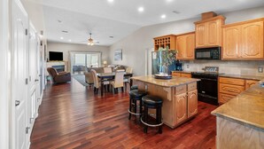 A spacious kitchen with wooden cabinets, a central island with stools, a dining area with a sofa, and a view to the living room. - Beach House - Hooked on the Cape by PHG (Port St. Joe)