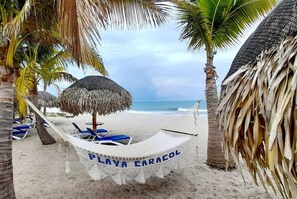Een hangmat met de tekst 'PLAYA CARACOL' op een zandstrand, met palmbomen en een rieten parasol.