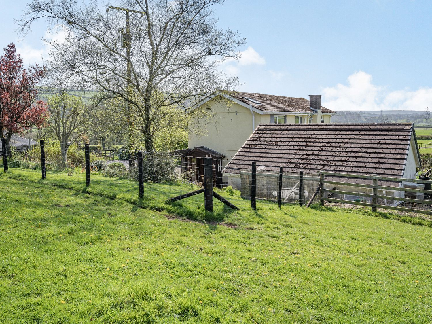 Una casa rural con techo inclinado, rodeada por un patio cercado con césped y árboles.