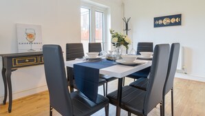 A dining area with a white table, black chairs, and a blue table runner. There is a window with blinds, a vase with flowers, and a wall-mounted artwork depicting the moon phases.