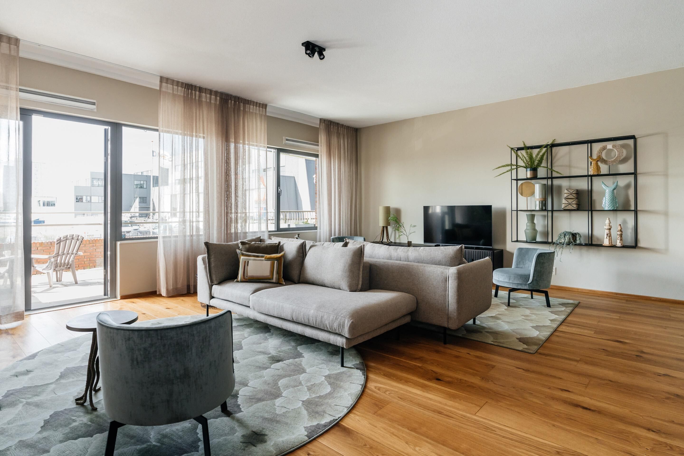 A modern living room with a grey sofa, a round coffee table, and a TV on a black stand. There is a large window with curtains, a balcony visible outside, and a wall-mounted shelf with decorative items.