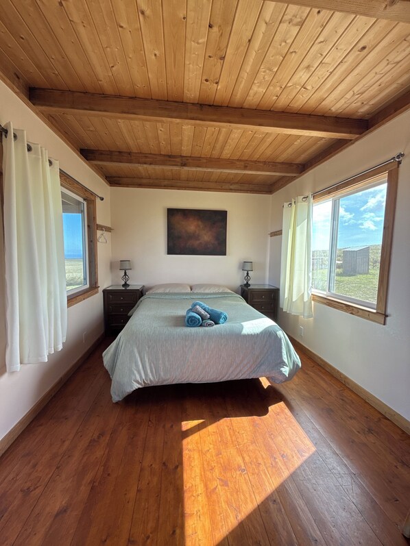Une chambre avec un plafond en bois, un lit recouvert d’une couverture grise, deux tables de chevet avec des lampes et des fenêtres avec des rideaux.