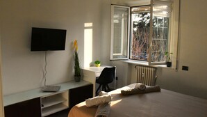 Une chambre d’hôtel avec un lit, un bureau avec un ordinateur, une télévision et une fenêtre donnant sur des arbres et une voiture.