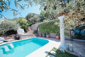 Una piscina con escalones de piedra y plantas en macetas, rodeada de olivos y un muro de piedra.