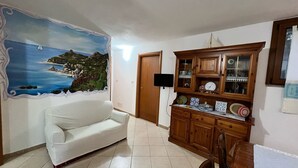 A living room with a white sofa, a wooden cabinet with glass doors, and a wall mural of a coastal scene. - Villa Eden 2' Sea + Free Parking (Sardegna)