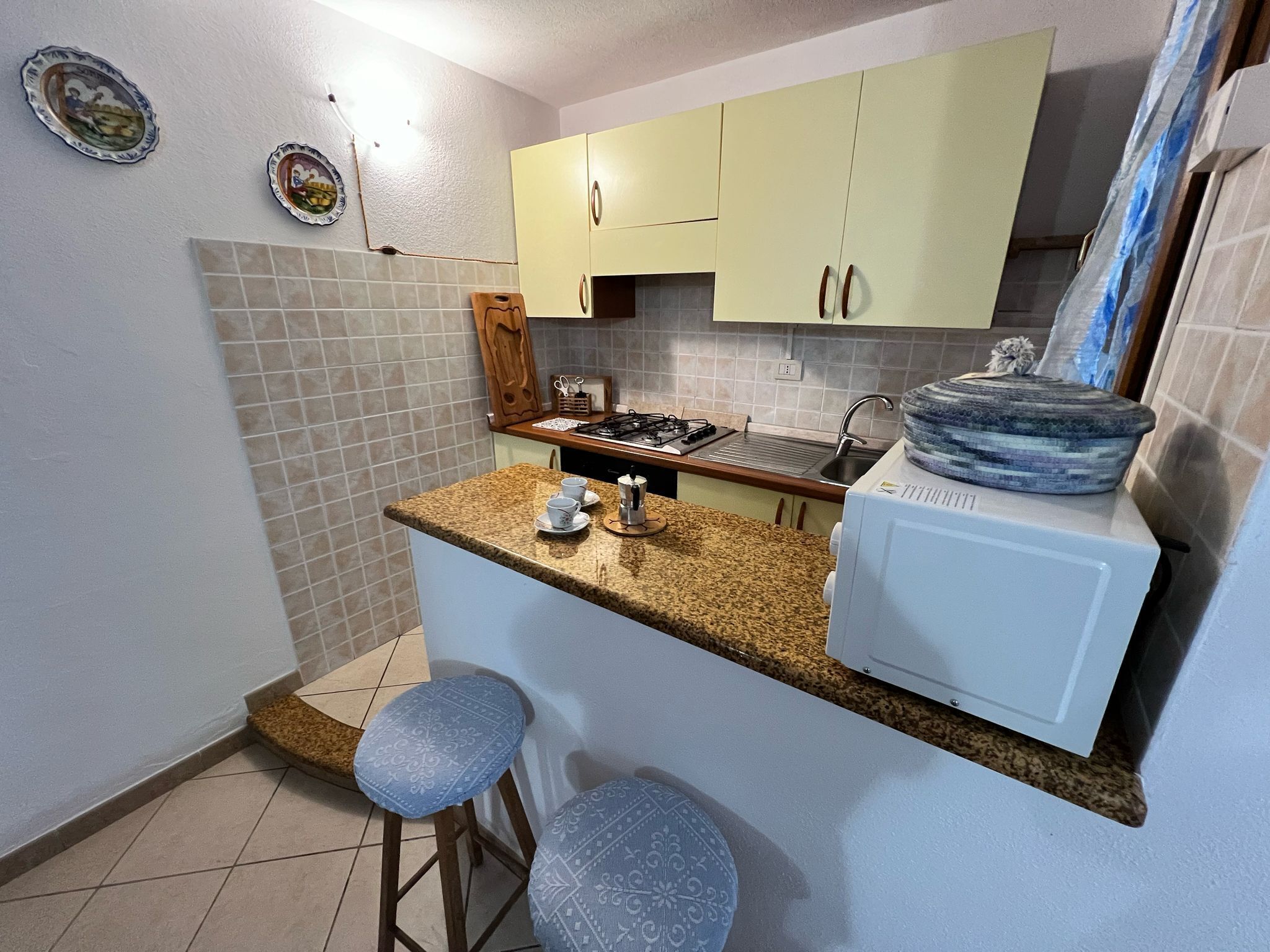 A compact kitchen with a gas stove, a sink, and a microwave. There are two stools and a small bar area.