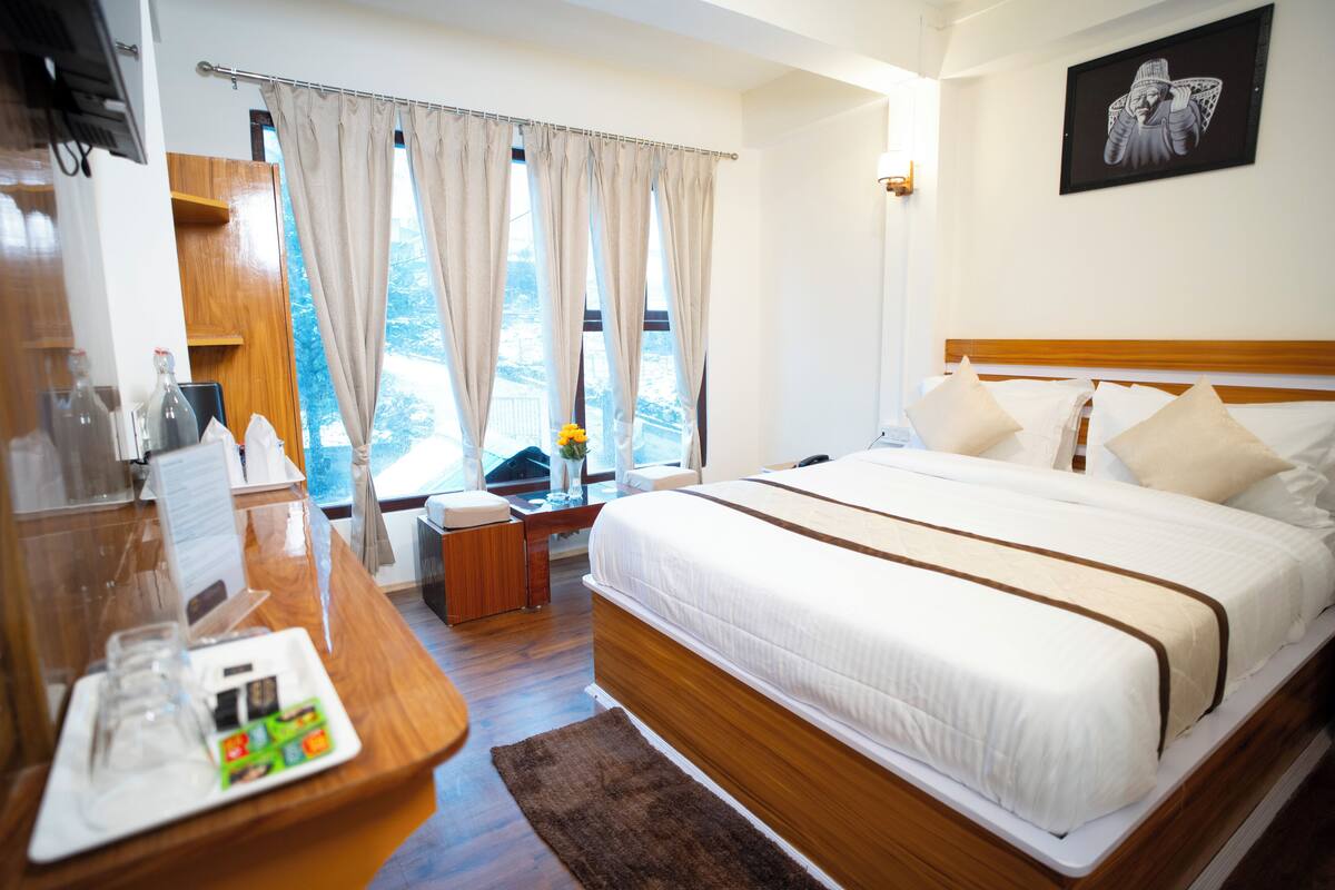 A hotel room with a large bed, a wooden desk with a glass of water, a small table with a vase, and a window with curtains.