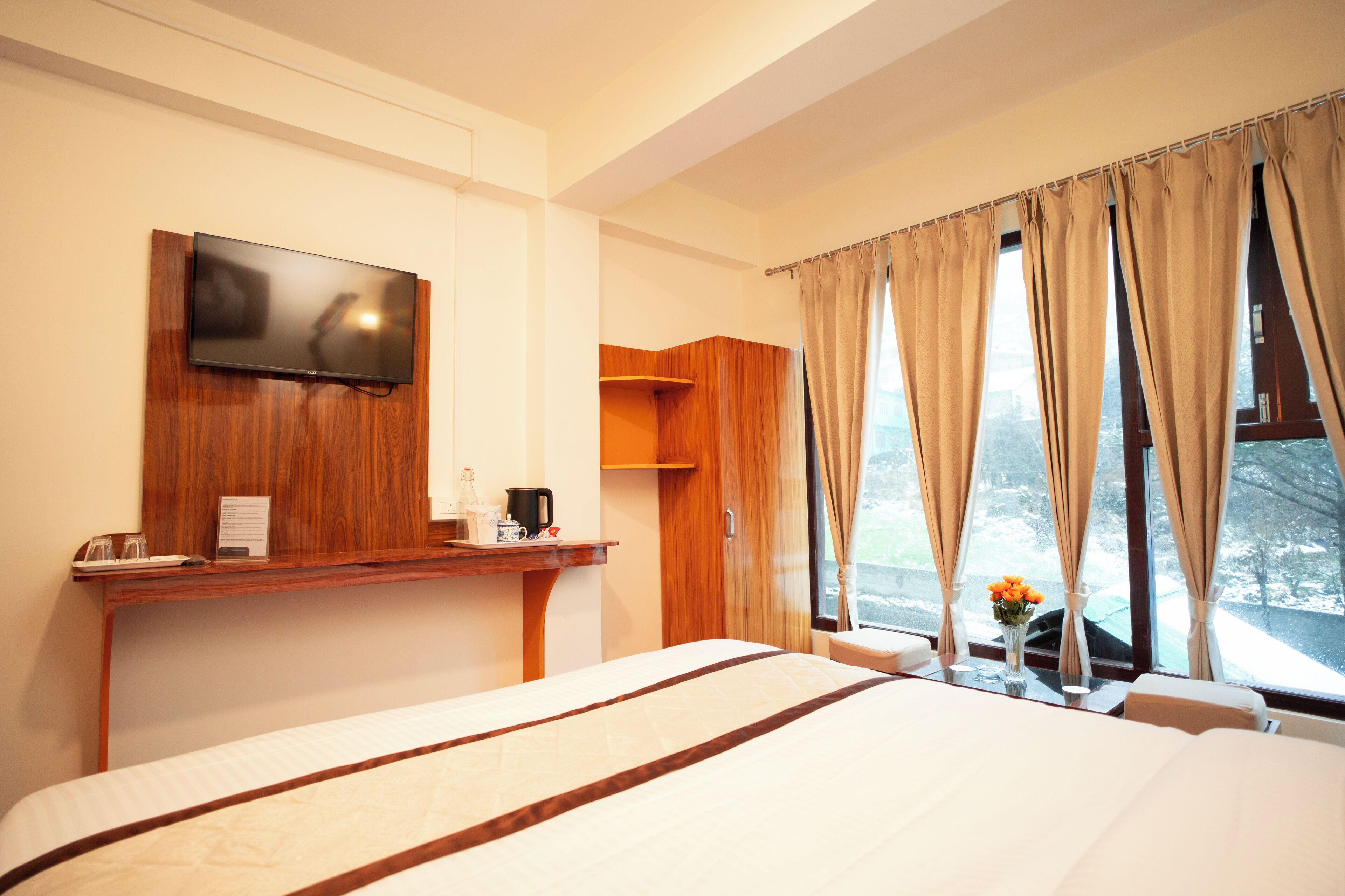 A hotel room with a large bed, a flat-screen TV mounted on a wooden panel, a small table with a coffee maker, and a window with curtains.