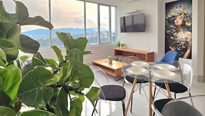 Un salon moderne doté d’une grande fenêtre, d’un téléviseur à écran plat, d’une table à manger avec des chaises et d’une grande plante.