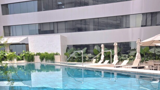 Une piscine intérieure moderne, avec des carreaux bleus et transparents, entourée de chaises longues et de parasols, et un bâtiment aux grandes fenêtres en arrière-plan.