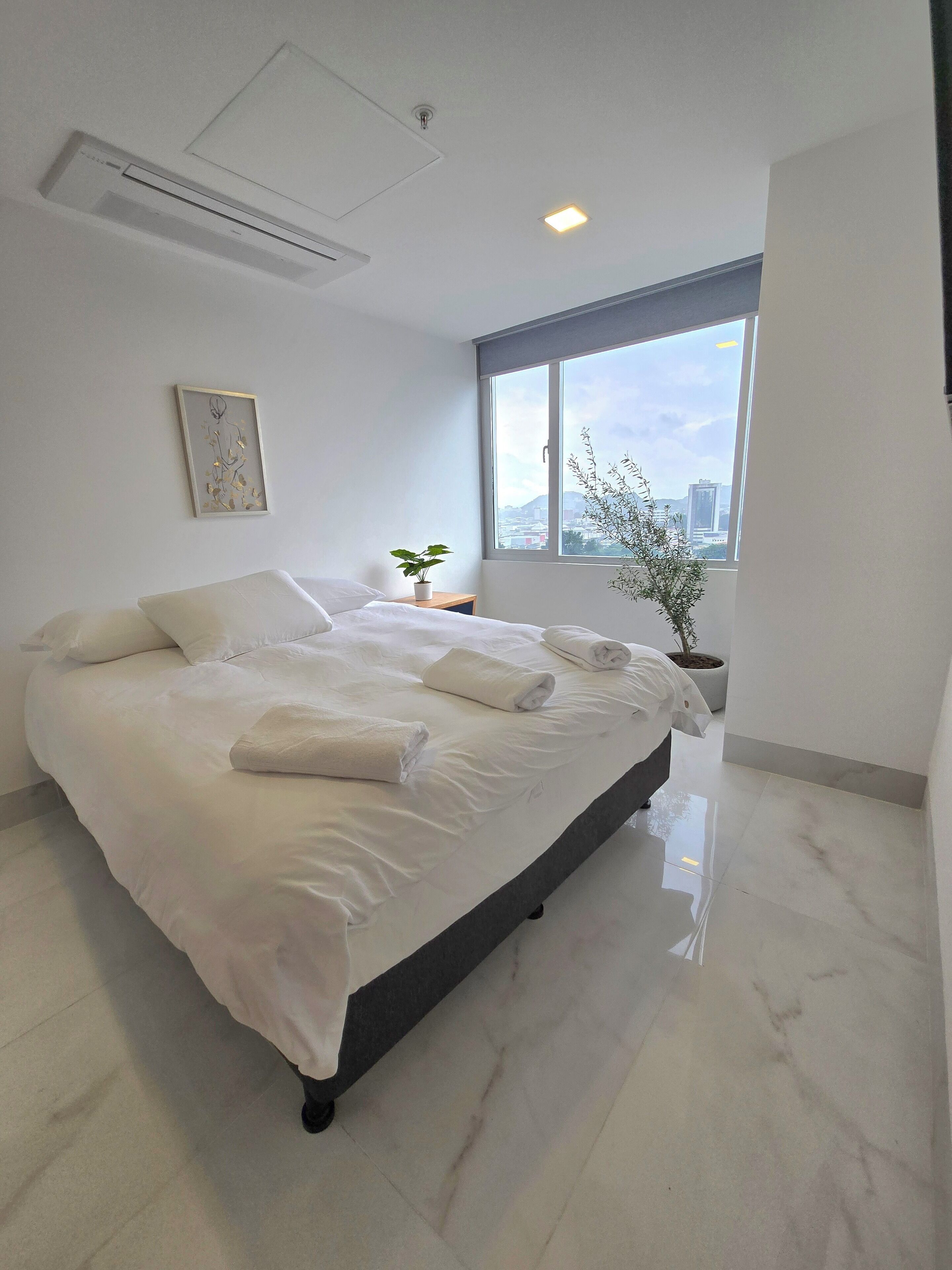 A modern bedroom with a large bed, a potted plant, and a city view through a large window.
