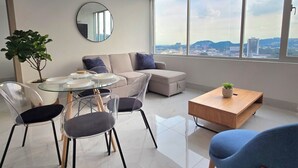 Un salon moderne avec une table à manger en verre, des chaises transparentes, un canapé et une table basse. Une grande fenêtre offre une vue sur la ville.