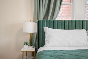Un lit bien fait, avec une tête de lit rembourrée de couleur verte, un oreiller blanc et une petite table de chevet sur laquelle se trouvent une lampe et un pot de fleurs.