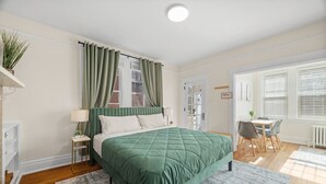 A bedroom with a bed, a nightstand, a lamp, a window with curtains, a radiator, and a dining area with chairs and a table.