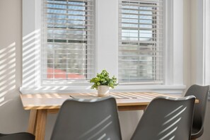 Un coin repas avec une table en bois et des chaises noires, une plante en pot posée sur la table et des stores blancs aux fenêtres.