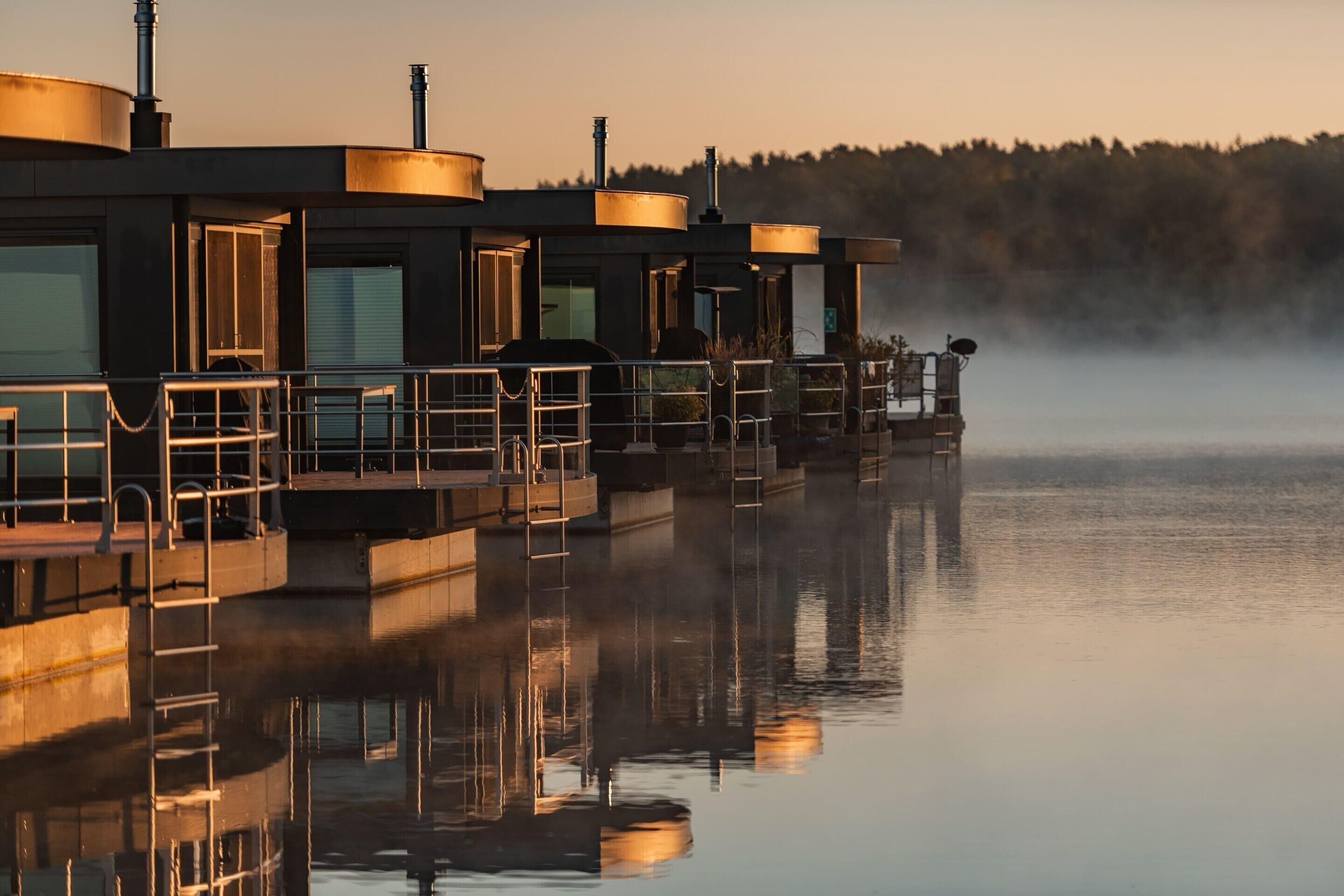 Un hôtel situé au bord de l’eau, composé de plusieurs bâtiments, doté d’espaces de détente en plein air et surplombant un plan d’eau calme dans lequel se reflètent les structures.