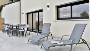 A modern outdoor seating area with a table and chairs, a sun lounger, and a large window with a view.